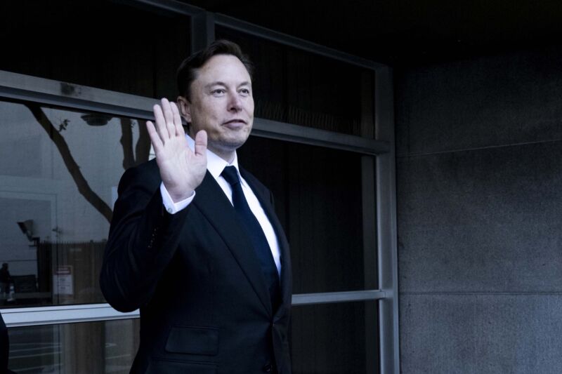 Elon Musk wearing a suit and waving with his hand as he walks away from a courthouse.
