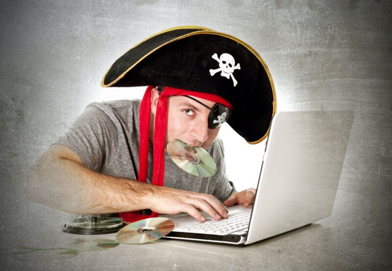 A man, surrounded by music CDs, uses a laptop while wearing a skull-and-crossbones pirate hat and holding one of the CDs in his mouth.