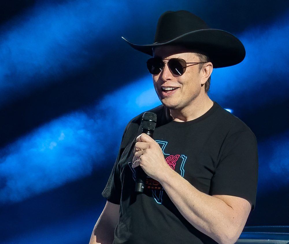 Elon Musk speaks at an event while wearing a cowboy hat, sunglasses, and T-shirt.