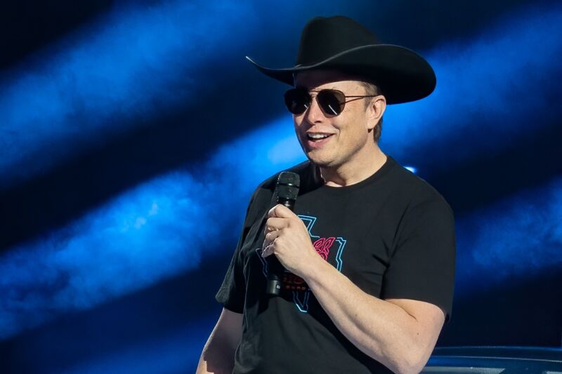 Elon Musk speaks at an event while wearing a cowboy hat, sunglasses, and T-shirt.