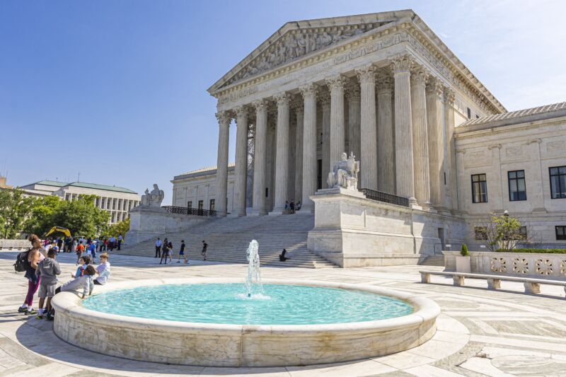 The US Supreme Court building is seen on a sunny day. Kids mingle around a small pool on the grounds in front of the building.