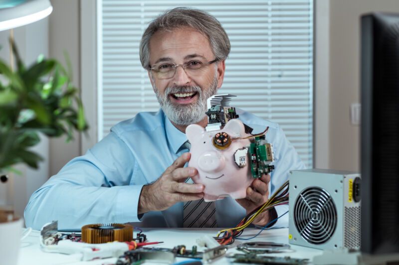 Man holding a piggy bank at his desk, with the piggy wired up with strange circuits and hardware