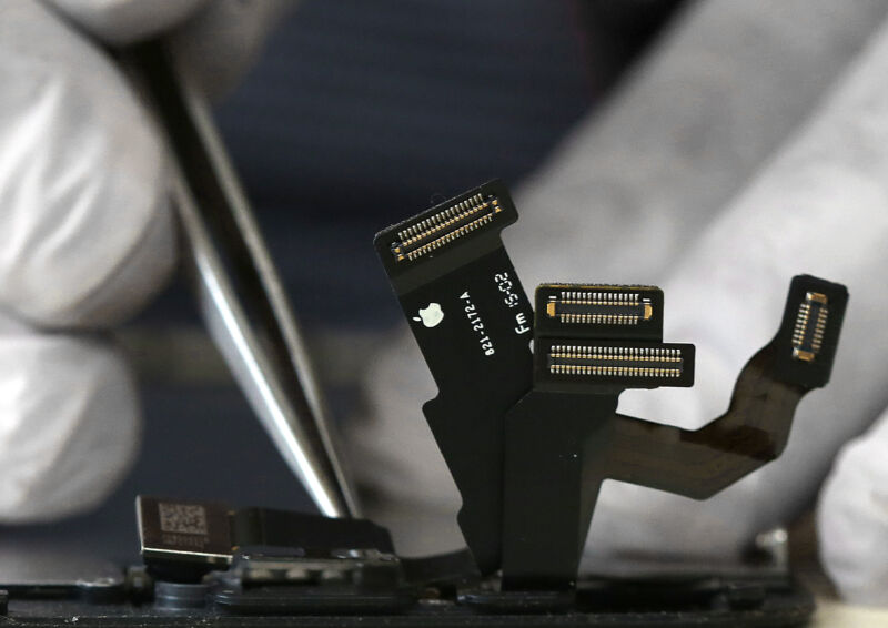 Cables que emanan de un iPhone en reparación, con las manos con guantes sosteniendo una pinza sobre el teléfono