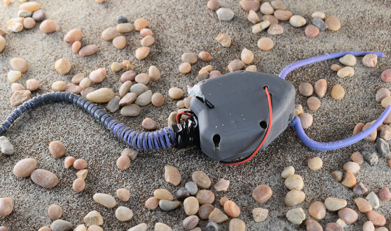 Image of a plastic robot with a triangular body and a long tail, perched on a sandy environment.