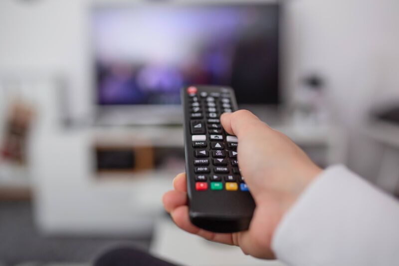 A person's hand aiming a cable TV remote control at a TV screen