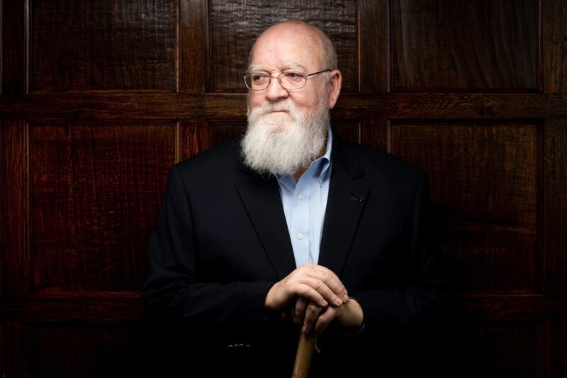 Daniel Dennett seated against black background in blue shirt, bowtie and dark jacket