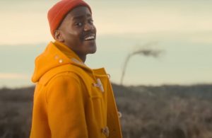 Gatwa in orange knitted cap and sweater, side view, smiling at the camera