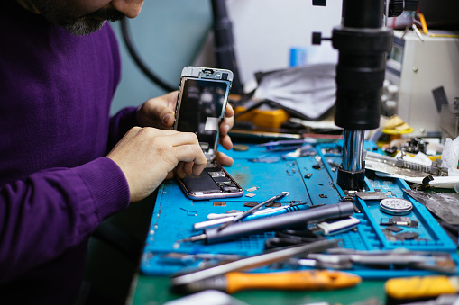 Technician repairing mobile phone at a station with microscope, heat gun, and blue mat