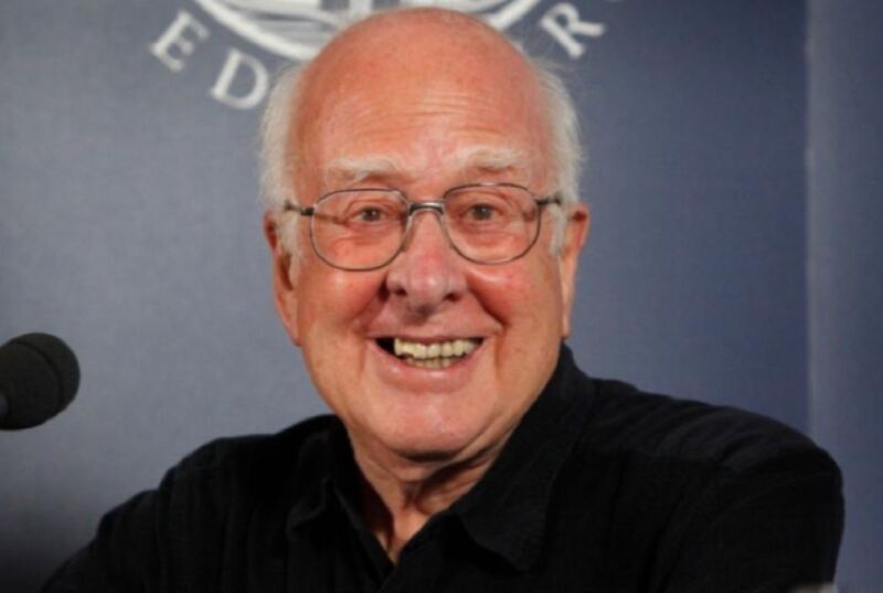 Smiling Peter Higgs, seated in front of microphone with Edinburgh logo in the background