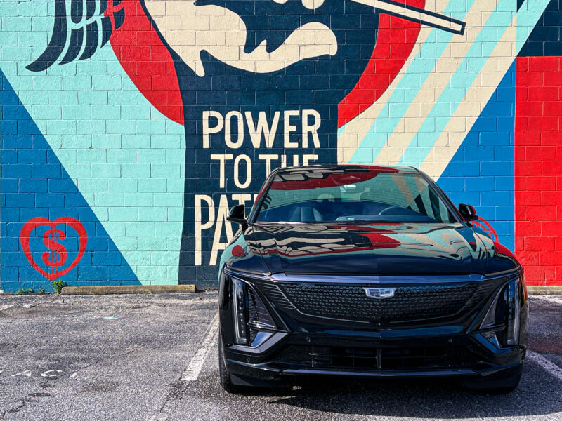 A black Cadillac Lyriq seen head-on, parked in front of a mural that says Power to the Patients