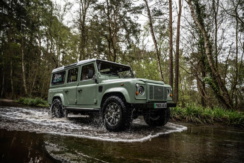 A green classic land rover defender drives through some standing water