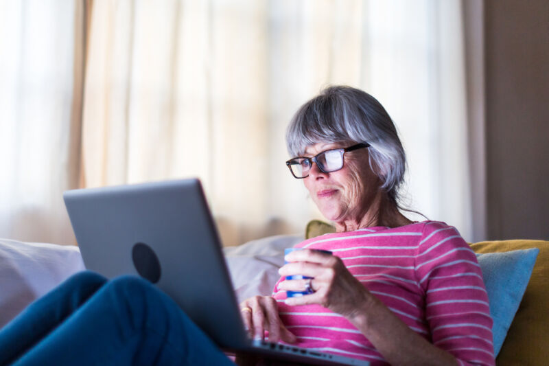 An older woman holding a coffee mug and staring at a laptop on her lap.