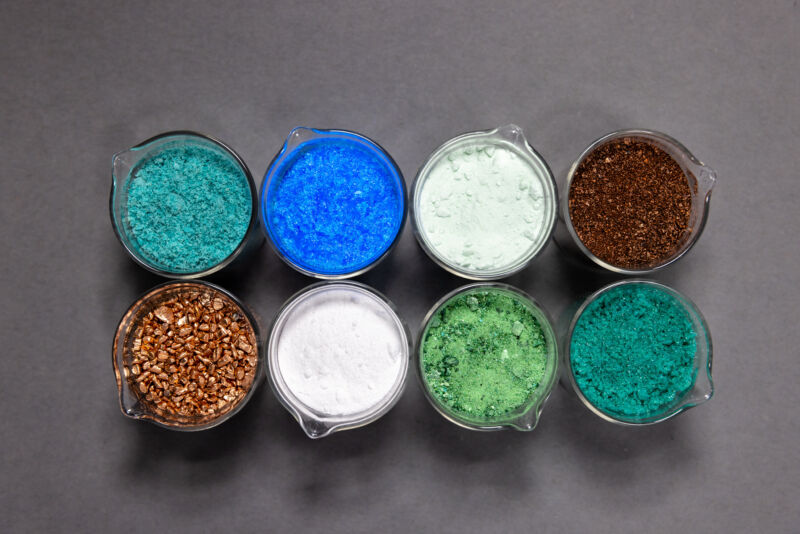 Eight beakers filled with colorful mineral salts, photographed from above.