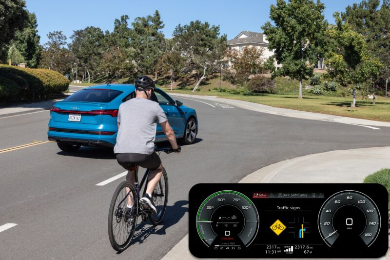 A cyclist rides near a blue Audi SUV. The image also shows a screenshot of the Audi dashboard, which has a large yellow icon warning the driver of a cyclist