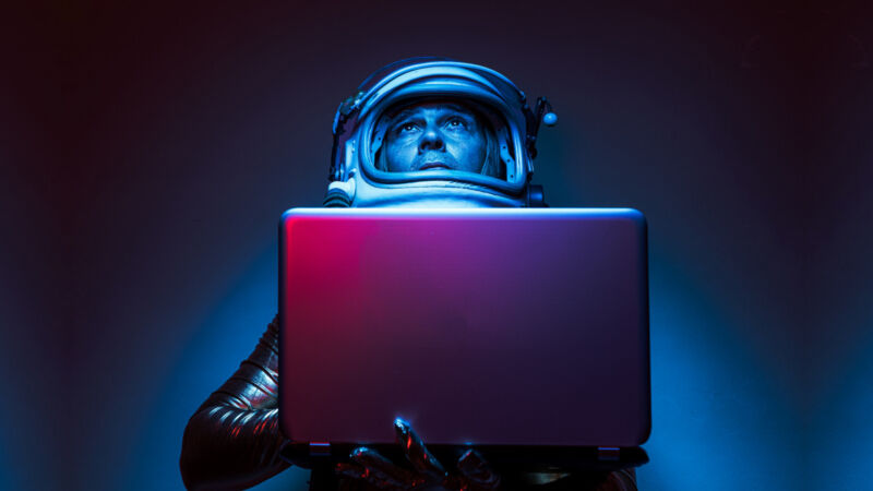 Close shot of Cosmonaut astronaut dressed in a gold jumpsuit and helmet, illuminated by blue and red lights, holding a laptop, looking up.