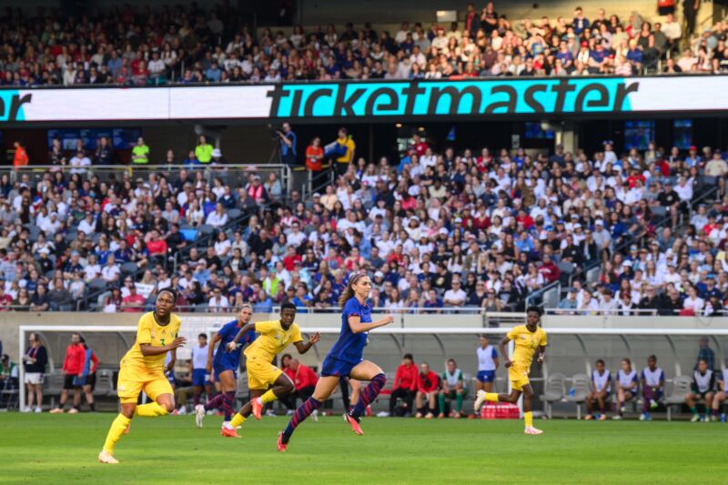 A large Ticketmaster logo is displayed on a digital screen above the field where a soccer game is played.