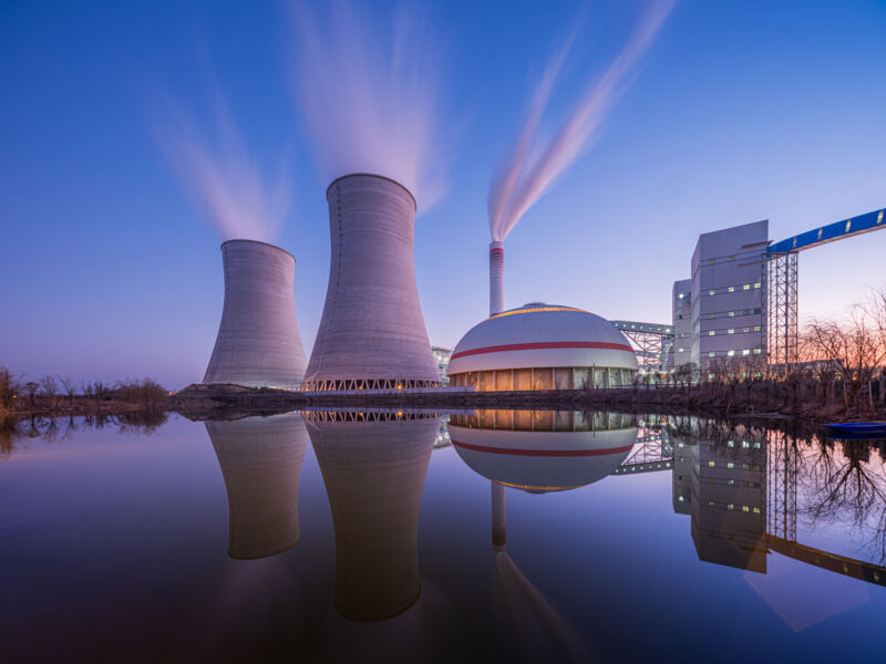 A nuclear reactor and two cooling towards on a body of water, with a late-evening glow in the sky.