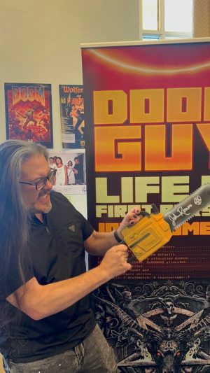 Romero poses with a toy chainsaw at an event promoting his autobiography, <a  data-cke-saved-href="https://romero.com/shop/p/doomguy" href="https://romero.com/shop/p/doomguy"><em>Doom Guy</em></a>.