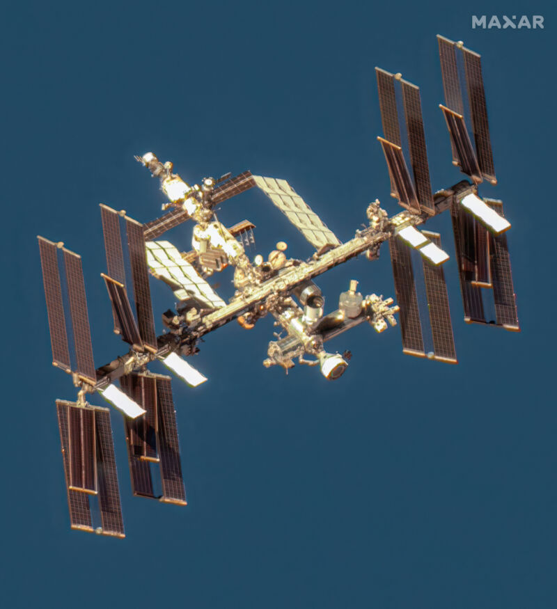 La Estación Espacial Internacional (ISS) en órbita alrededor de la Tierra, capturada desde el espacio. La estructura está compuesta por varios módulos y grandes paneles solares que se extienden a ambos lados. La ISS aparece iluminada por la luz solar, destacándose sobre un fondo oscuro que sugiere el espacio exterior.