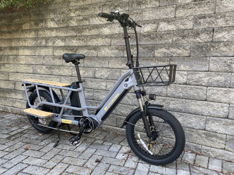 Image of a grey, long-tail cargo bike with tall handlebars and a basket in front, and a wooden deck and footrests by the cargo area.