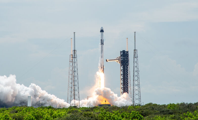 Un cohete Falcon 9 de SpaceX despegando desde la plataforma de lanzamiento en Cabo Cañaveral, Florida. La nave espacial Crew Dragon se encuentra en la parte superior del cohete, transportando astronautas en una misión hacia la Estación Espacial Internacional. En el fondo, se observan estructuras de soporte y una columna de humo generada por los motores durante el lanzamiento.