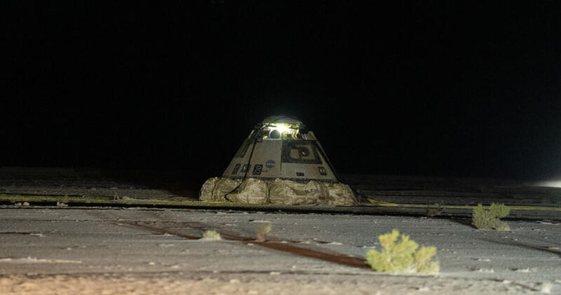 La cápsula espacial Starliner de Boeing, recién aterrizada en el desierto de Nuevo México durante la noche. La nave está iluminada y se puede ver apoyada sobre airbags desplegados, que amortiguaron su descenso. El fondo es oscuro, resaltando la cápsula contra el paisaje desértico.