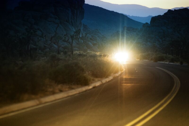 Blinding bright lights from a car pierce through the dark scene of a curved desert road at dusk. The lights form a star shaped glare. Double yellow lines on the paved road arc into the foreground. Mountains are visible in the distant background.