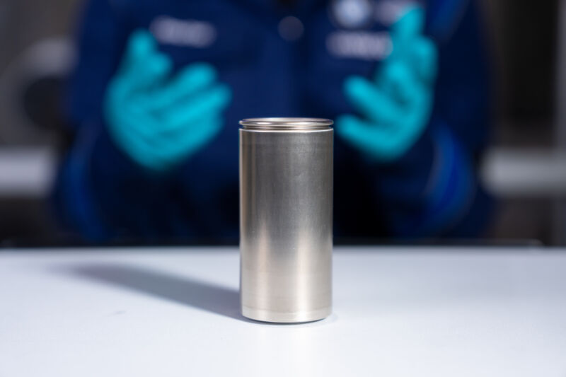 in the foreground, a large silver cylindrical battery cell. In the background, a pair of nitrile glove-clad hands