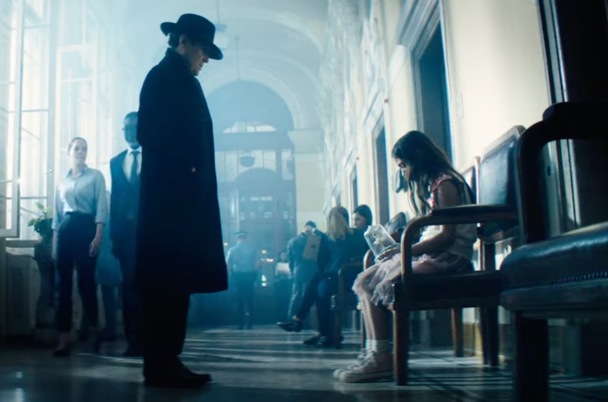 standing man in hat in a train station in front of a young sad girl sitting on a bench