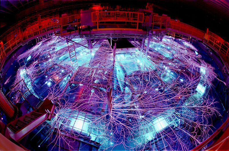 Image of a large, circular chamber covered filled with a lot of mechanical equipment, all of which is lit by blue internal glows and covered with massive, branching trails of lightning.