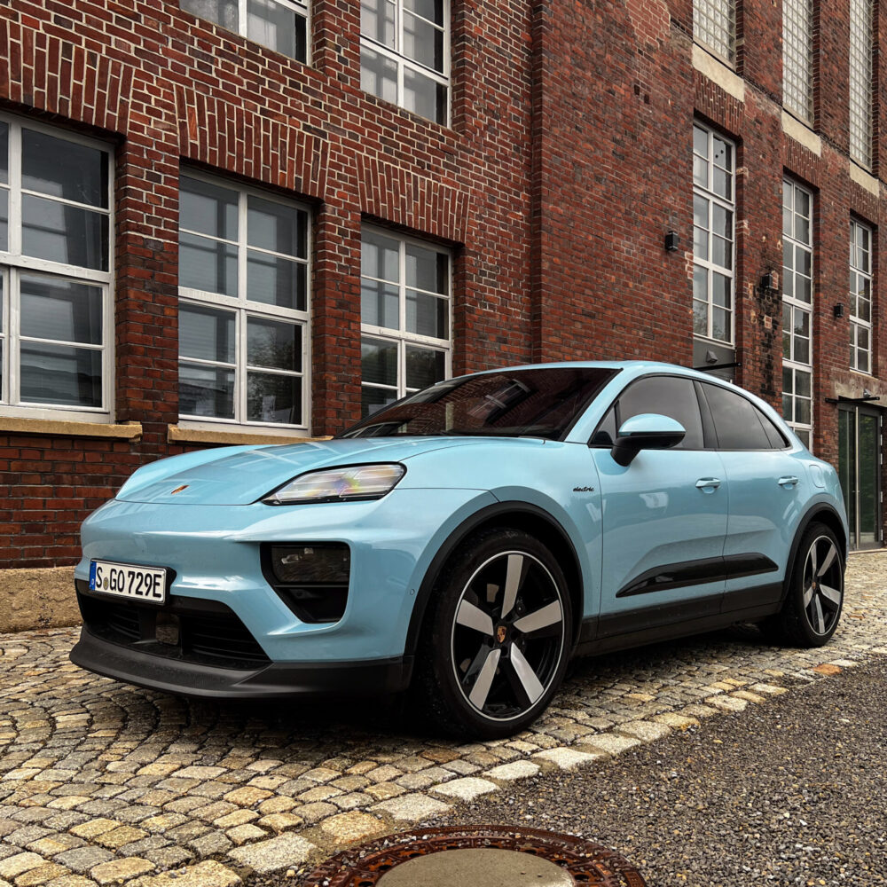 A pale blue Porsche Macan EV next to an old German industrial building.