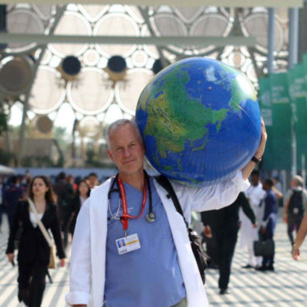 A person wearing a lab coat walks through a crowd, carrying a large world globe on his shoulder.