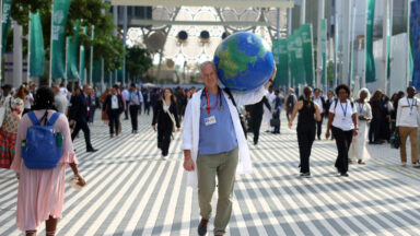 A person wearing a lab coat walks through a crowd, carrying a large world globe on his shoulder.