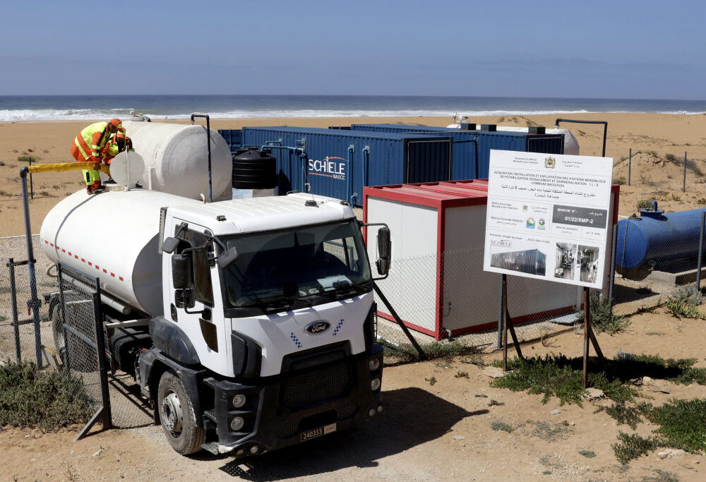 Image of a small tanker truck parked next to a few shipping container shaped structures, which are connected by pipes to storage tanks.