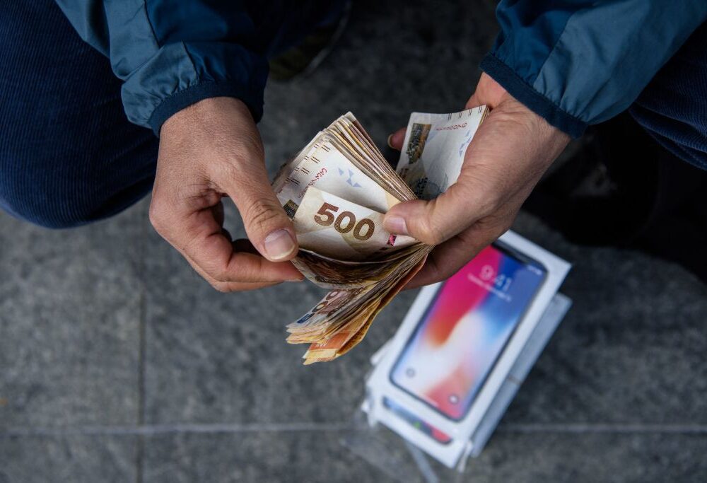 Una persona contando una cantidad significativa de billetes, incluyendo uno de 500 euros en primer plano. En el suelo, junto a sus manos, se puede ver una caja de un iPhone, lo que sugiere una posible transacción relacionada con dispositivos electrónicos. La imagen evoca la idea de una compra o intercambio de dinero en efectivo por productos tecnológicos.