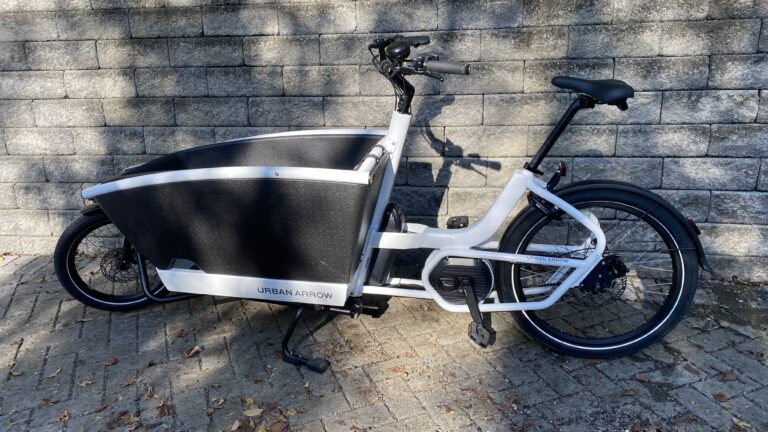 Image of a front-loading cargo bike with white metal tubes, set against stone pavement and walls.