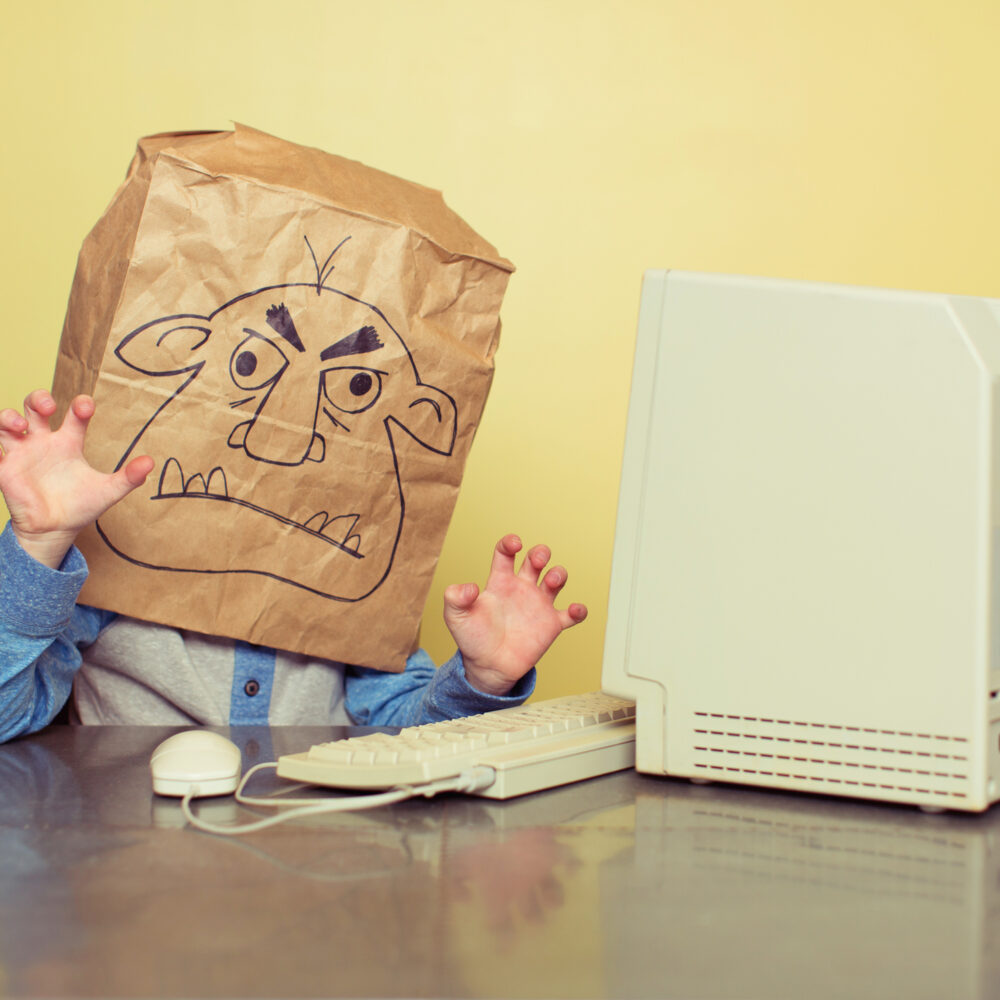 Picture of a person using an old Mac with a paper bag over his head. The bag has the face of a troll drawn on it.