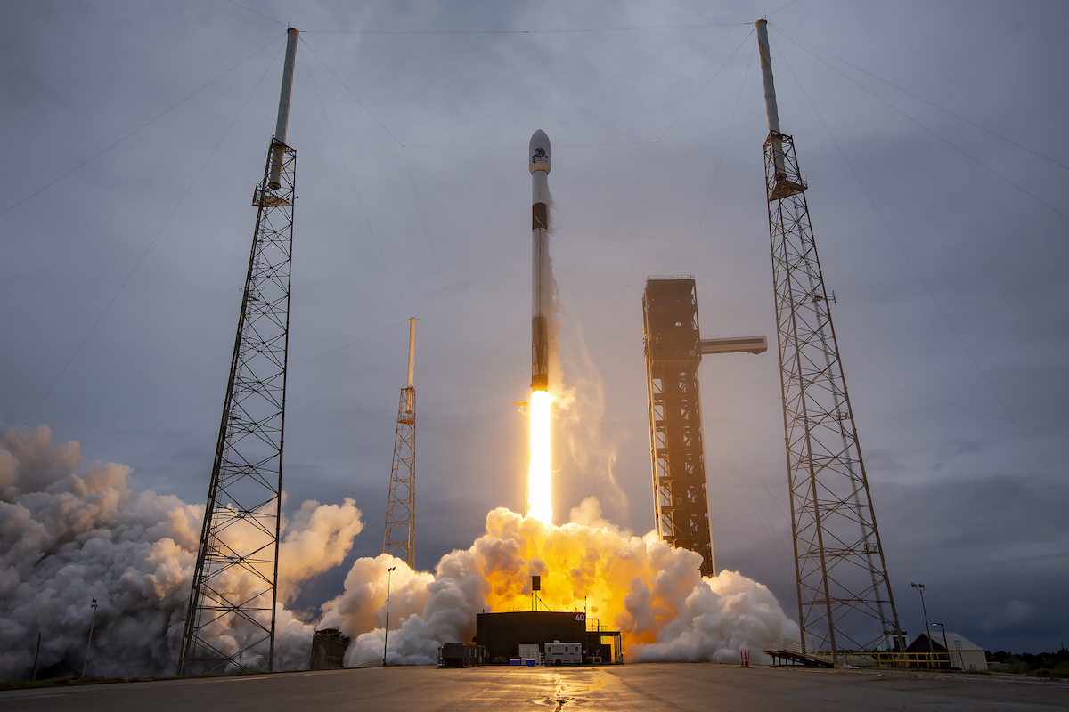 Lanzamiento de un cohete Falcon 9 de SpaceX desde la estación espacial de Cabo Cañaveral, Florida, con un cielo nublado y humo saliendo de la base mientras el cohete se eleva. El Falcon 9 está en plena fase de despegue, impulsado por sus motores mientras deja una estela de fuego y humo en su trayectoria ascendente.