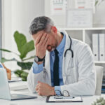 A frustrated doctor sitting at a desk facepalming himself.