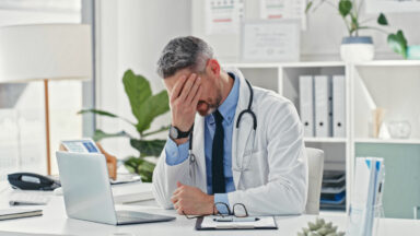 A frustrated doctor sitting at a desk facepalming himself.