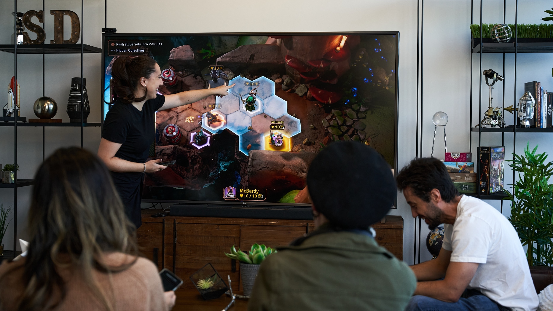 Friends gathered in front of a living room TV, with one person standing up and pointing at a hex on the game grid.