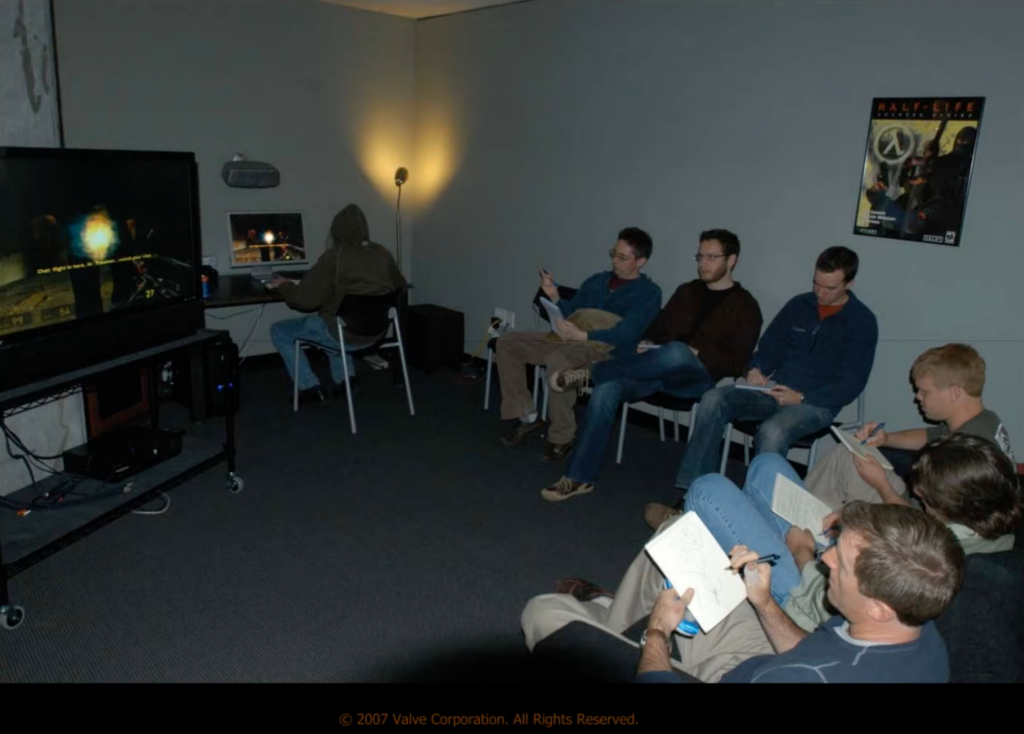 A group of game developers sits on couches and takes notes while a PC gamer plays Half-Life 2