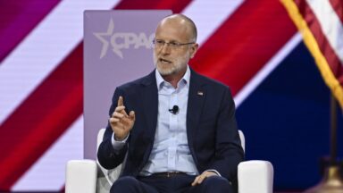 Federal Communications Commission member Brendan Carr sits on a stage and speaks while gesturing with his hand. Behind him is the CPAC logo for the Conservative Political Action Conference.