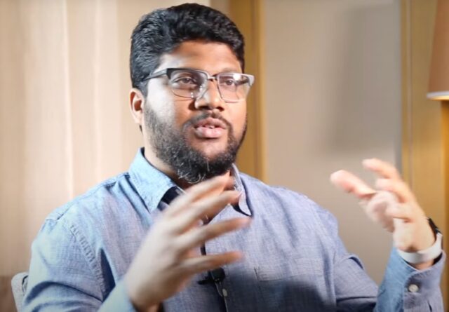 Indian man with beard in blue dress shirt gesturing as he talks
