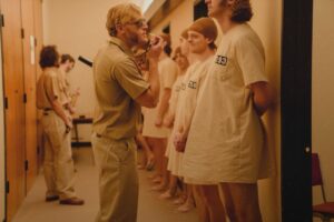 Blond man dressed as prison guard wearing sunglasses, harassing prisoners in hospital gowns.