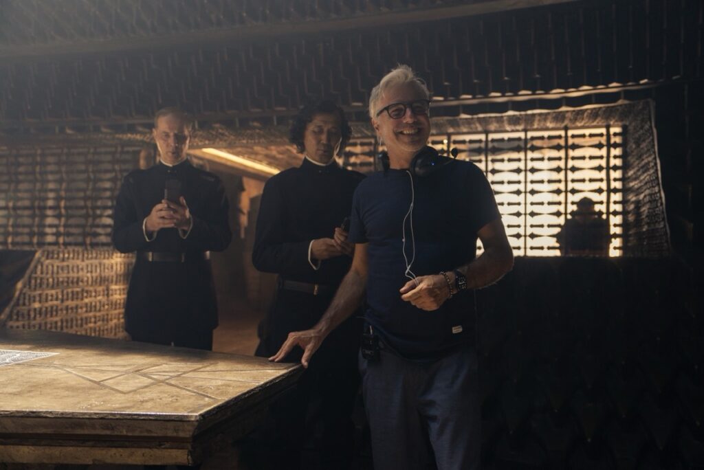 smiling white haired man in glasses, jeans and t-shirt on set with other crew members