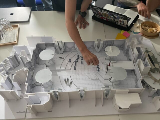 a white model of a set with someone's arm placing miniature people inside it.