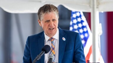 Rhode Island Gov. Dan McKee speaks during a news conference from behind a podium and two microphones, with an American flag in the background.