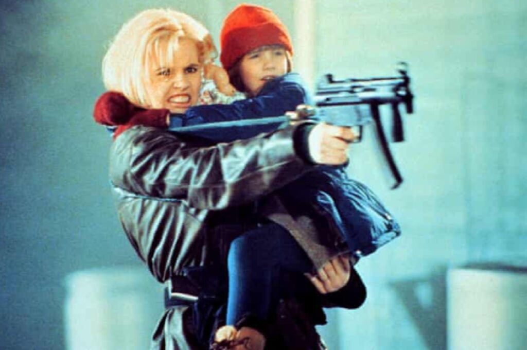 Blonde woman holding a young girl while shooting an automatic weapon