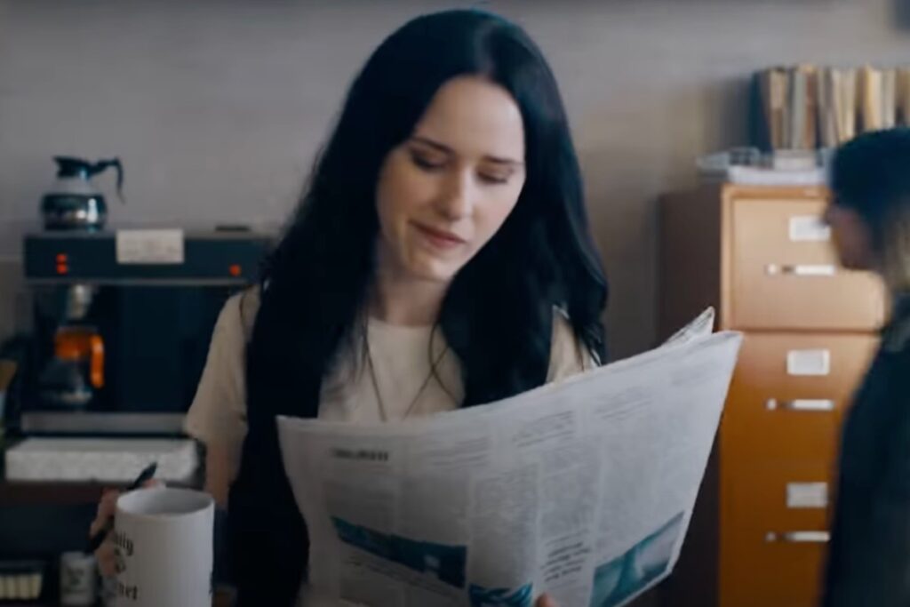 dark haired woman reading a newspaper while standing with a cup of coffee in her hand.